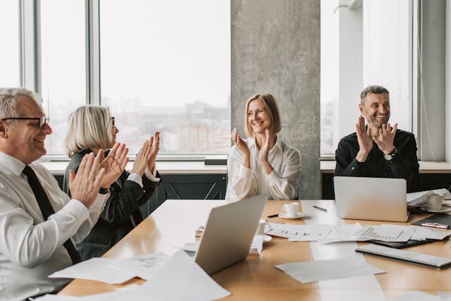 Zwei Frauen und zwei Männer unterschiedlichen Alters in förmlicher Bürokleidung sitzen gemeinsam an einem Tisch, auf dem psychologische Unterlagen ausgebreitet sind. Sie applaudieren zufrieden lächelnd.