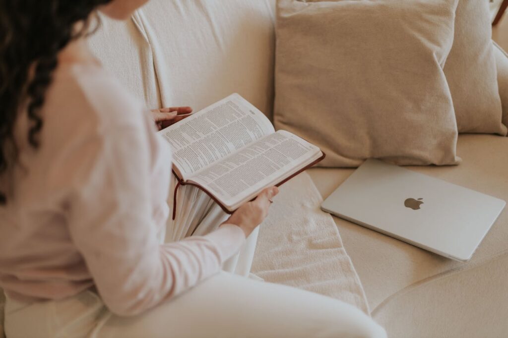 Eine Frau sitzt bequem auf dem Sofa und hält nachdenklich ein offenes Buch in beiden Händen, dessen Fachrichtung nicht erkennbar ist. Vor ihr liegt ein geschlossener Laptop.
